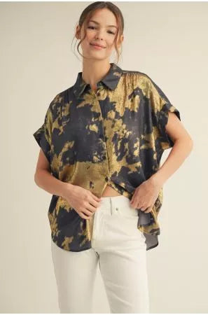 A woman modeling a short-sleeve button-up shirt with a black and gold abstract pattern, paired with white pants, against a plain light beige background.