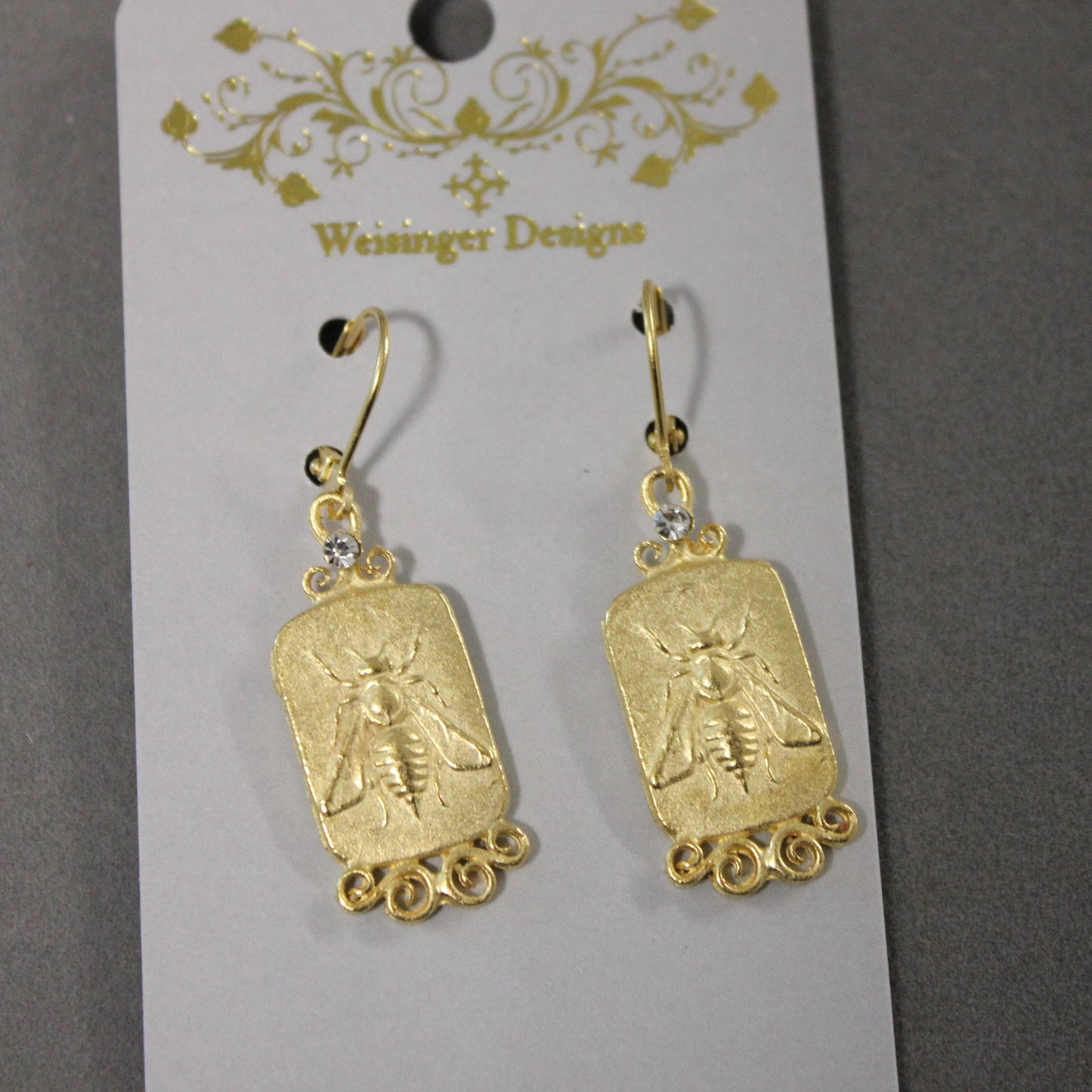 A pair of gold-toned drop earrings with rectangular bee-shaped charms and small clear rhinestones, hanging from a white display card printed with the brand name 'Weisinger Designs'.