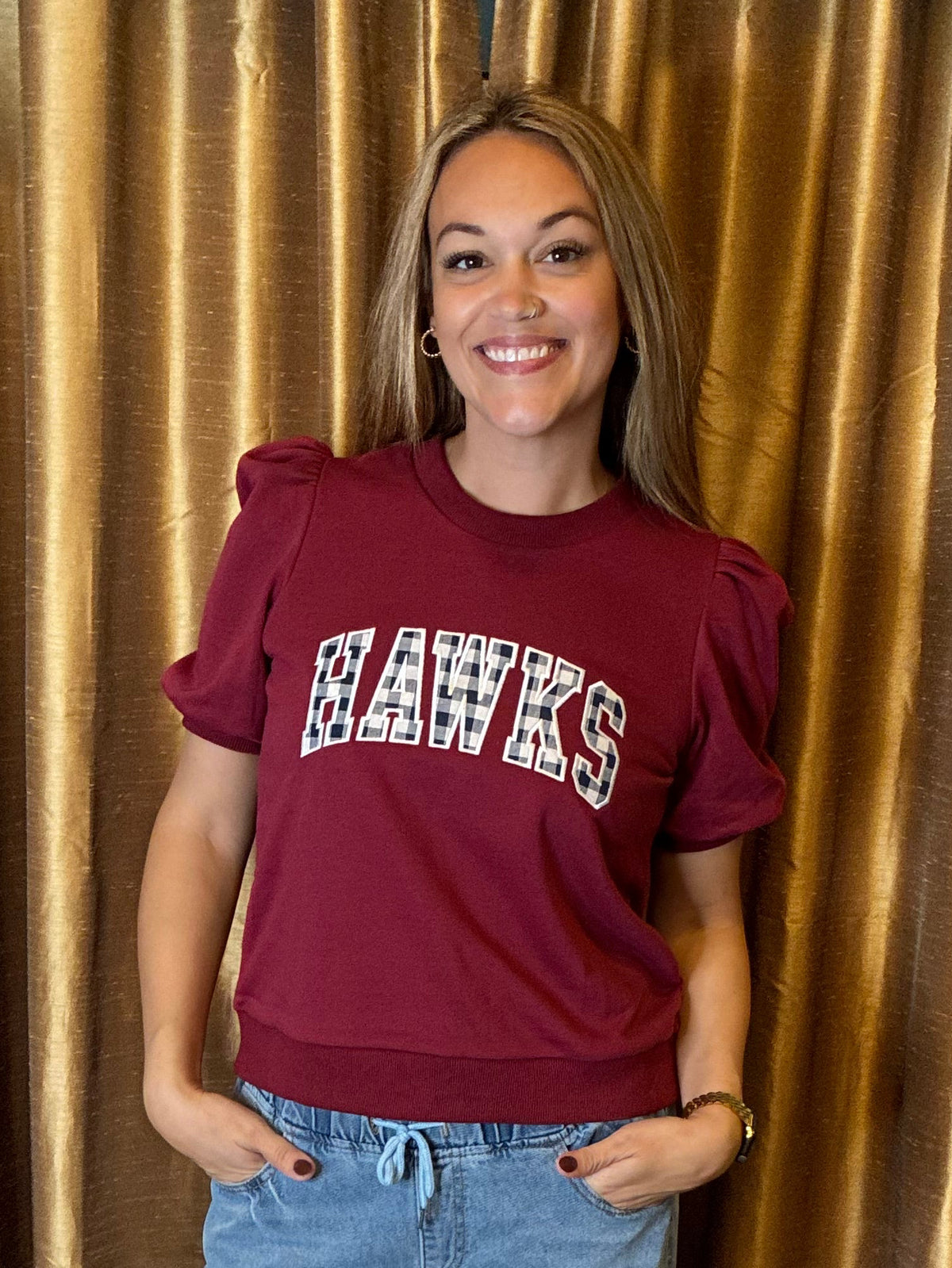 A smiling woman wearing a maroon short-sleeve sweatshirt with 'HAWKS' embroidery sew on across the chest in white outlined letters of navy plaid , paired with light blue jeans, standing in front of a gold curtain backdrop.