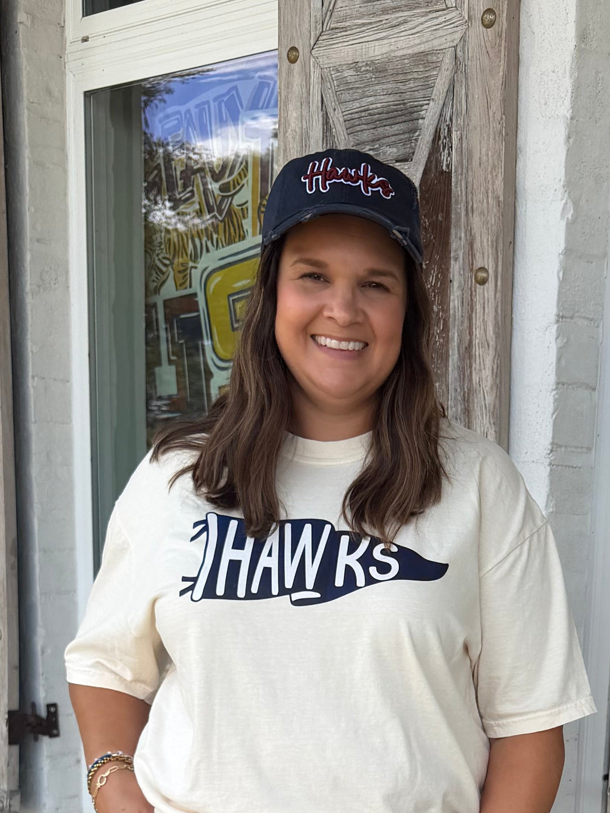 A woman wearing a cream graphic t-shirt with 'HAWKS' printed on the front, light blue flared jeans, and a Hawks navy baseball cap.