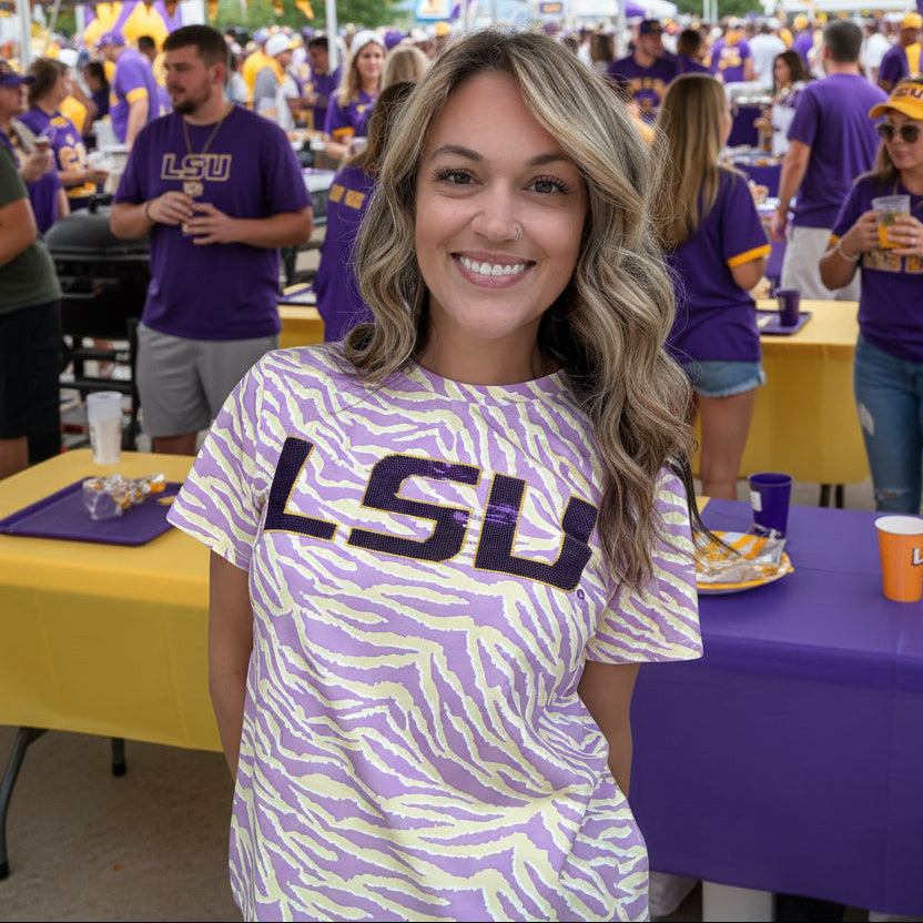 A smiling woman with blonde-highlighted hair wearing a purple and yellow tiger-print t-shirt with 'LSU' printed across the chest,