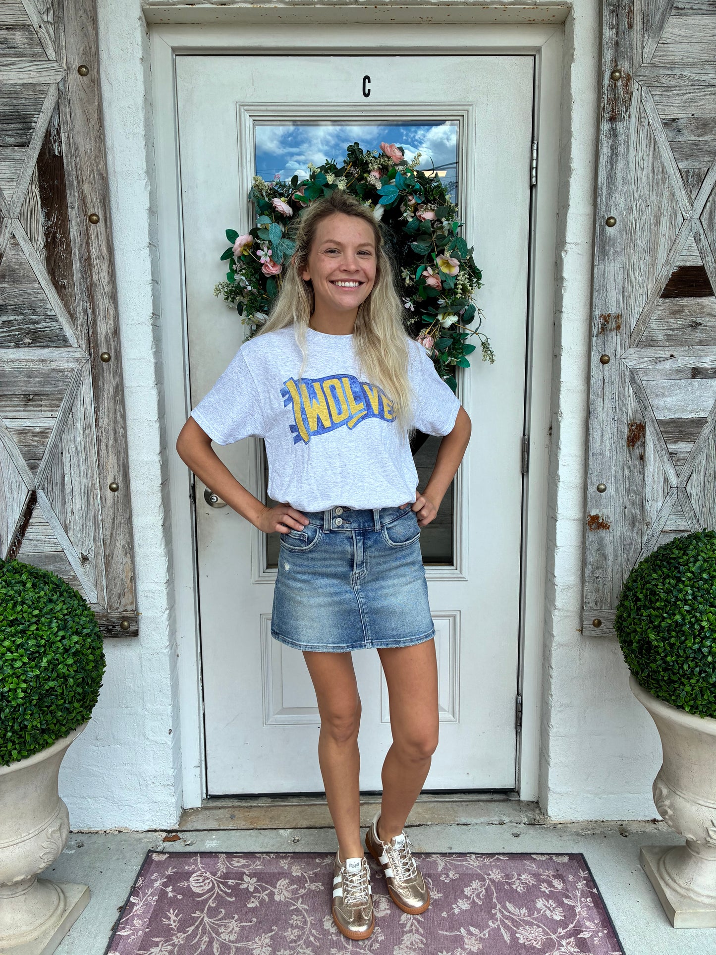 A young woman stands smiling on a front porch wearing an ash grey t-shirt with a blue and yellow wolves flag pennant ' design, a denim mini skirt, and metallic gold sneakers. The background features a white door with a green wreath, weathered wooden shutters, and potted greenery.