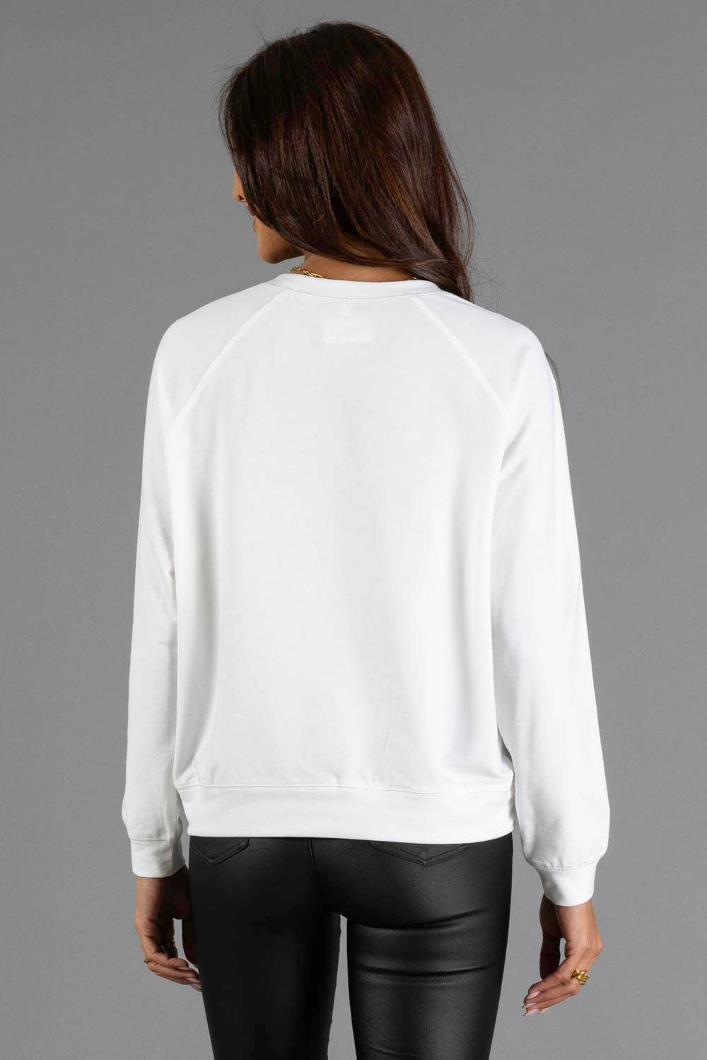 A woman viewed from the back wearing a white long-sleeve crewneck sweatshirt and black pants, set against a solid gray studio background.