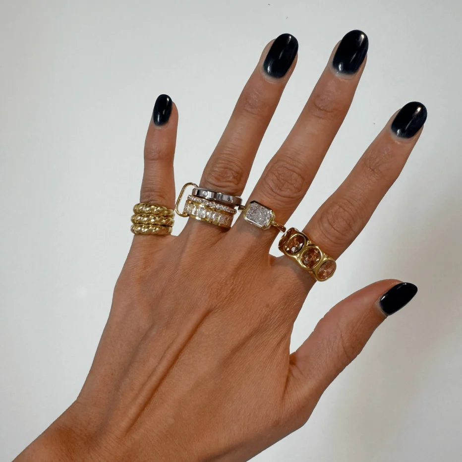 A close-up of a hand with black manicured nails wearing five different rings, including gold, silver, and white gold bands with clear stones, set against a plain off-white background.