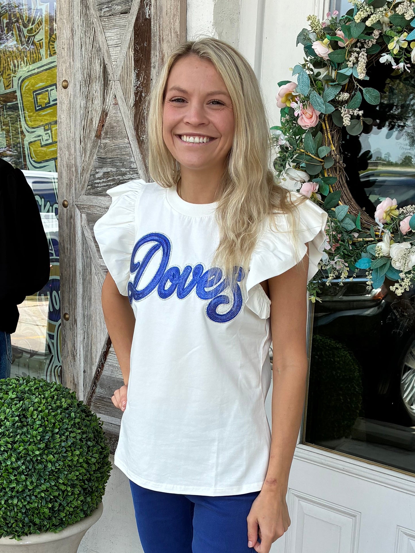 Woman wearing Doves Flutter Sleeve White Tee with blue sequin patch and silver border, ruffled flutter sleeves.