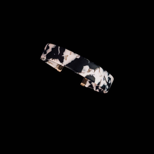 A slim rectangular cuff bracelet with a black and white marbled pattern and small gold-toned metal accents, shown on a solid black background.
