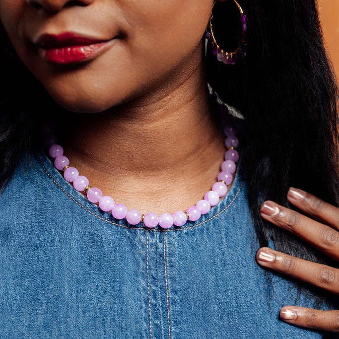A close-up lifestyle shot of a woman wearing a light purple/lavender beaded necklace and matching purple hoop earrings, paired with a blue denim top.