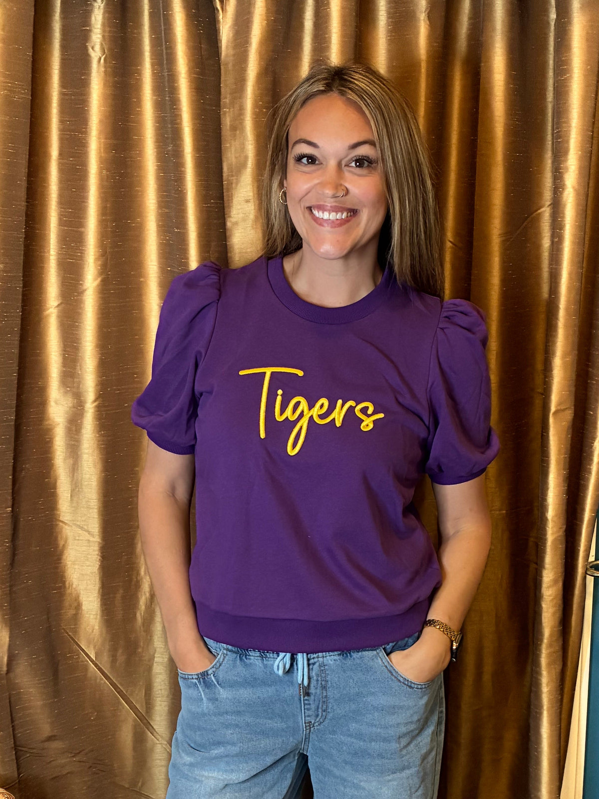 A woman stands indoors wearing a purple puff  short-sleeve shirt with 'Tigers' puff embroidery in yellow script across the chest, paired with light blue ripped jeans and black sandals. She is posed in front of a gold curtain and blue wall.