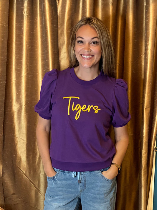 A woman stands indoors wearing a purple puff  short-sleeve shirt with 'Tigers' puff embroidery in yellow script across the chest, paired with light blue ripped jeans and black sandals. She is posed in front of a gold curtain and blue wall.