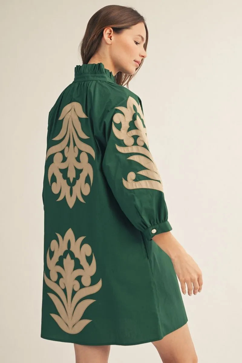 A woman wearing a dark green shift dress with large beige damask-style embroidery on the back and sleeve, shown from a side-back angle against a plain light beige studio background.