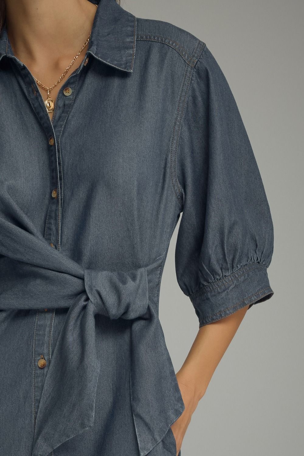 Close-up of a person wearing a blue chambray button-up shirt dress with puff sleeves and a tied waist, shown against a plain gray studio background.