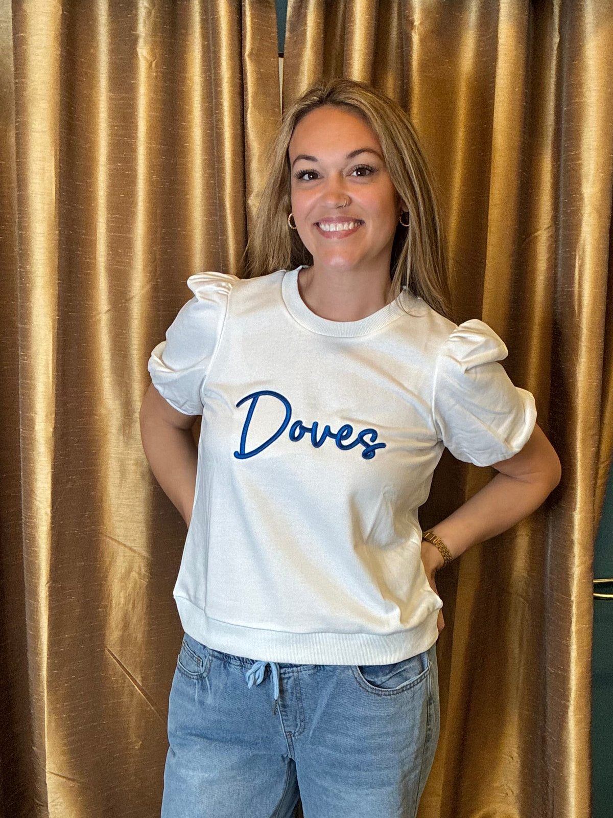 A woman stands indoors wearing a white short-sleeve t-shirt with 'Doves' printed in blue script across the chest, paired with light-wash ripped jeans and sandals. She is posed in front of a gold curtain with a blue wall behind it.