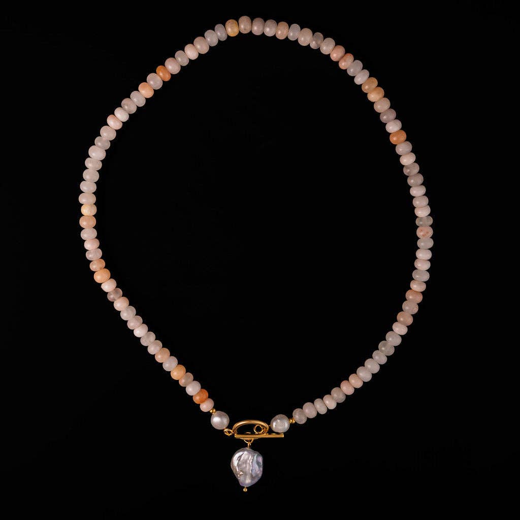 A beaded necklace made of small round beads in soft peach, cream, and pale pink tones, with a gold-tone toggle clasp and a large irregular pearl pendant, displayed on a solid black background.