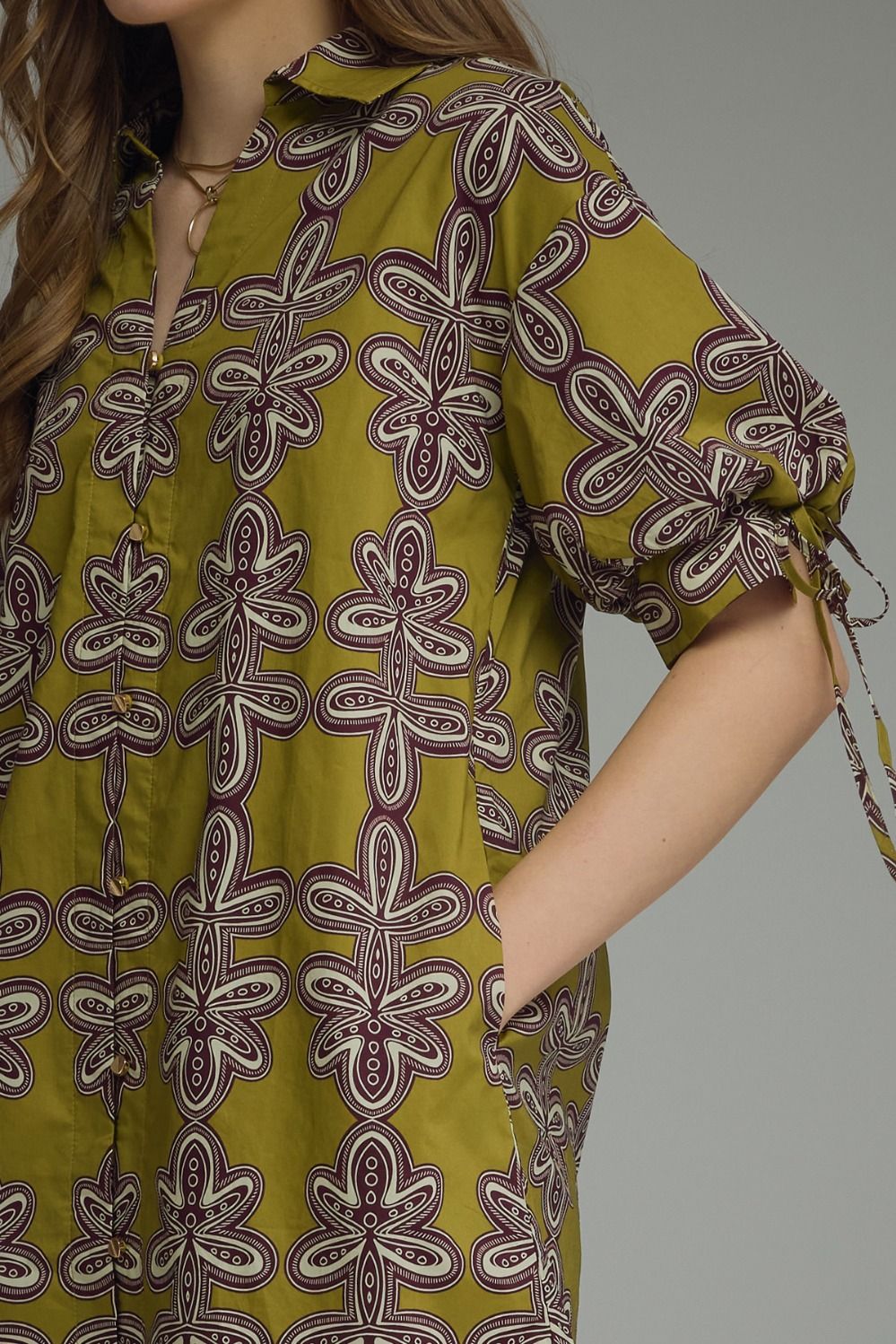A close-up of a woman wearing a short-sleeve button-front shirt dress in olive green with a large purple and white floral pattern. The dress has tie details on the sleeve cuffs and side pockets.