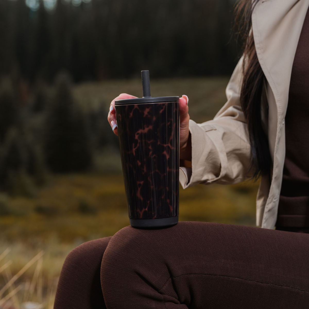 Person seated outdoors holding a Mocha Tortoise HydroJug Everyday Tumbler with a dark lid and straw