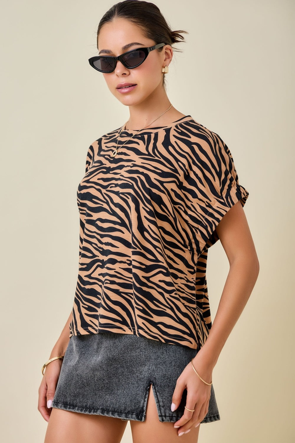 A woman wearing a short-sleeve crewneck top with a tan and black tiger print, styled with a gray denim skirt and black sunglasses against a solid beige background.
