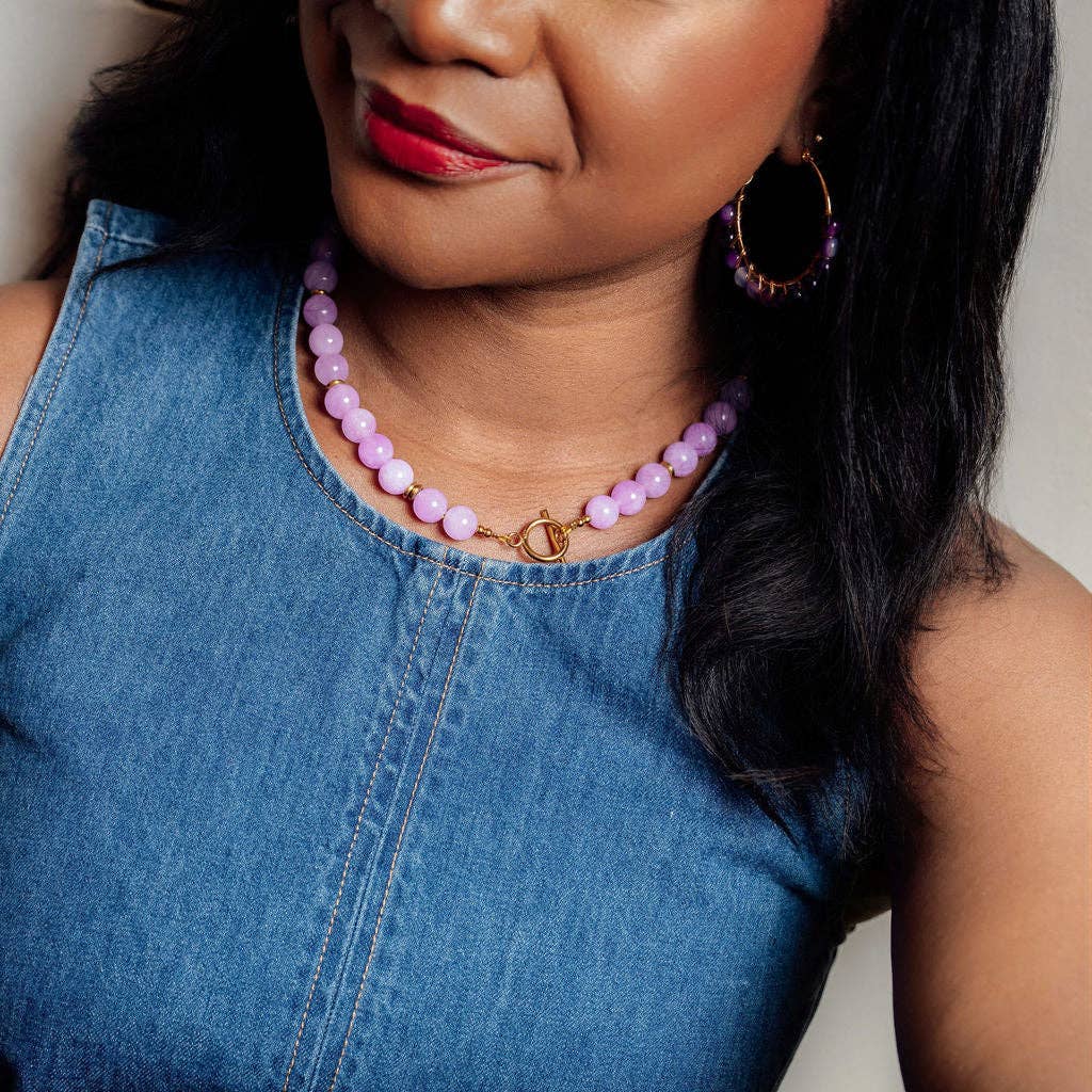 A close-up of a woman wearing a light purple beaded necklace with a gold clasp and matching purple hoop earrings, paired with a blue denim top.