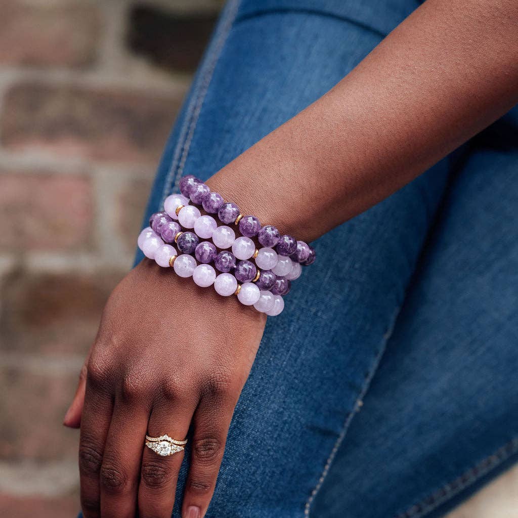 A close-up of a person's wrist wearing three stacked beaded bracelets made of purple and white marbled beads, paired with blue jeans and a gold ring.