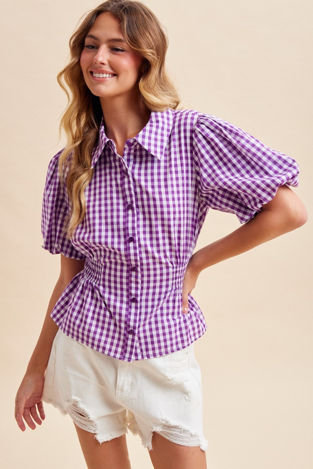 A woman wearing a purple and white gingham short-sleeve blouse with puff sleeves and a peplum waist, paired with white distressed shorts, against a solid beige background.
