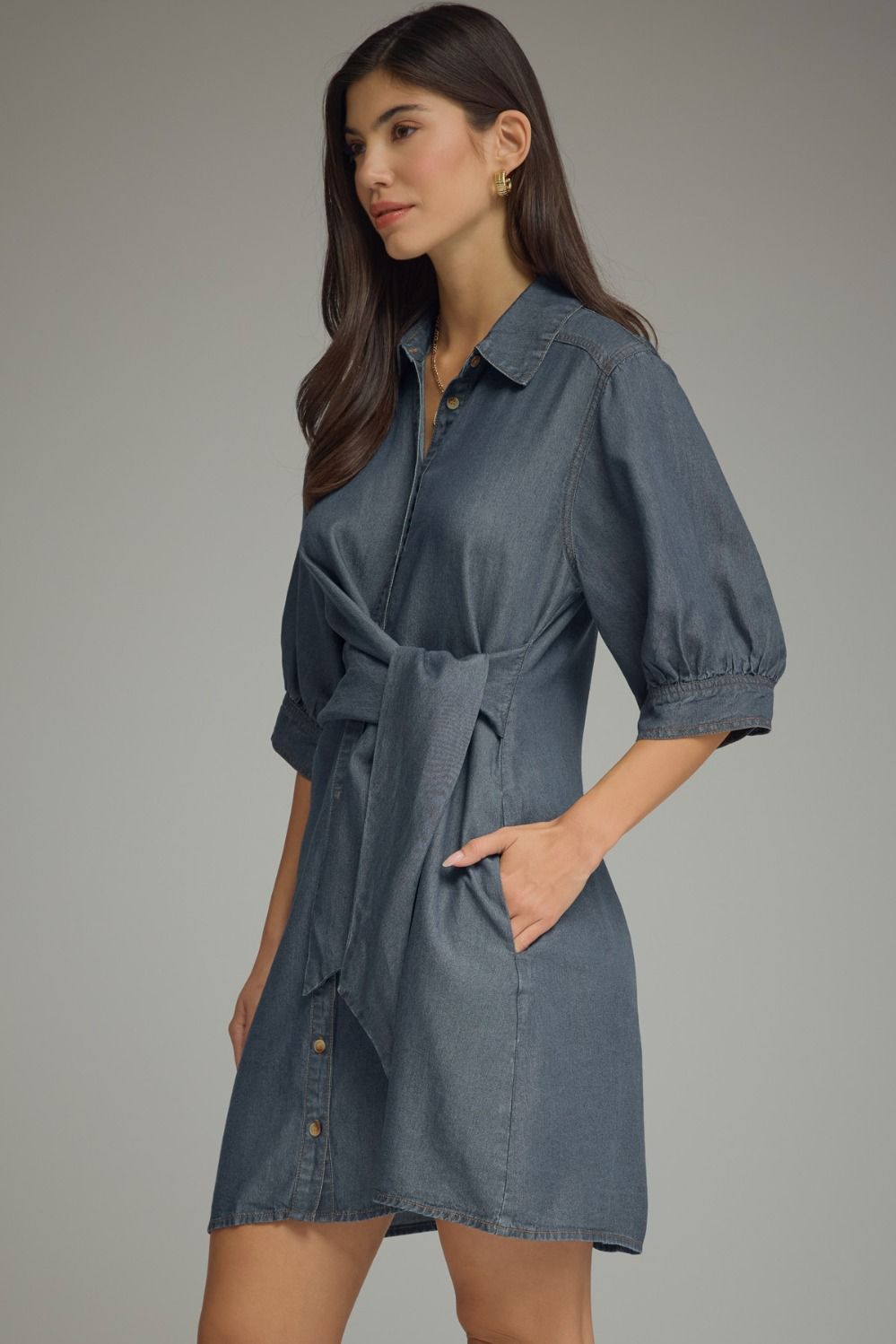 A woman modeling a slate blue denim shirt dress with puff sleeves, a self-tie belt, and button details on the front, set against a solid gray studio background.