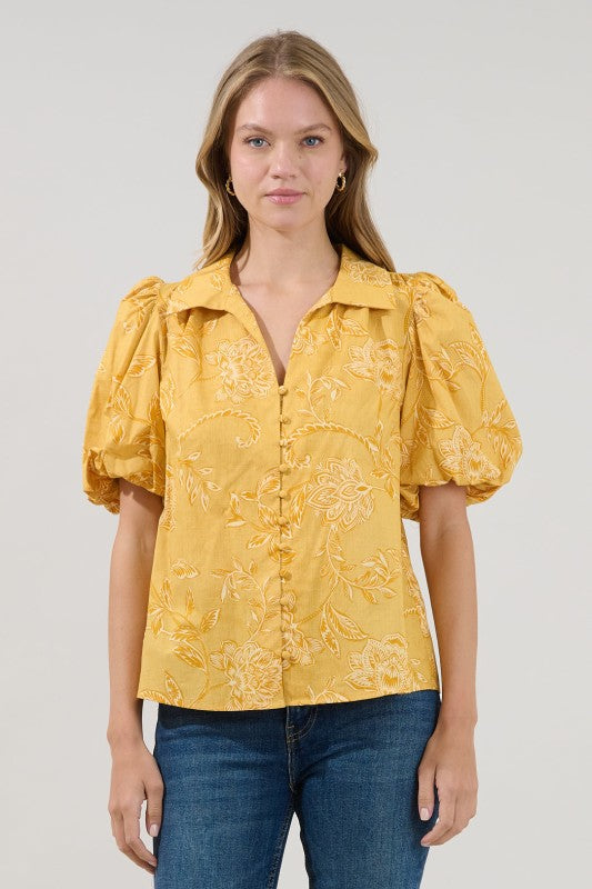 A woman modeling a yellow short-sleeve blouse with puff sleeves and a white floral jacquard pattern, paired with blue jeans, against a light gray studio background.