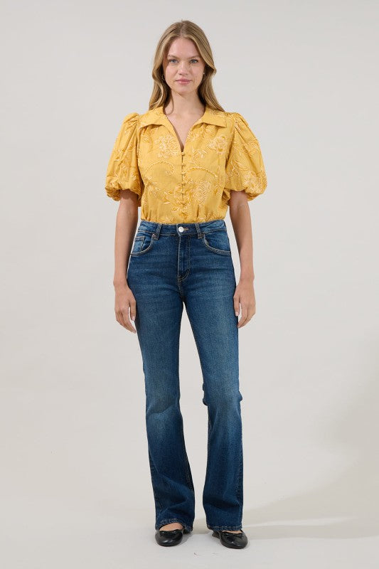A woman modeling a yellow short-sleeve blouse with textured puff sleeves and blue high-waisted flare jeans, standing against a light gray studio background.