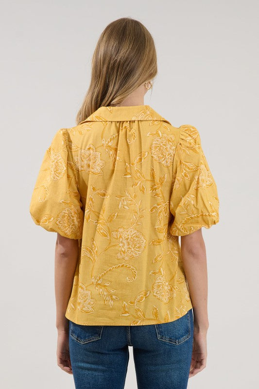 A woman viewed from the back wearing a mustard yellow short-sleeve blouse with a tonal floral jacquard pattern and puffed sleeves, paired with blue jeans, against a light gray studio background.