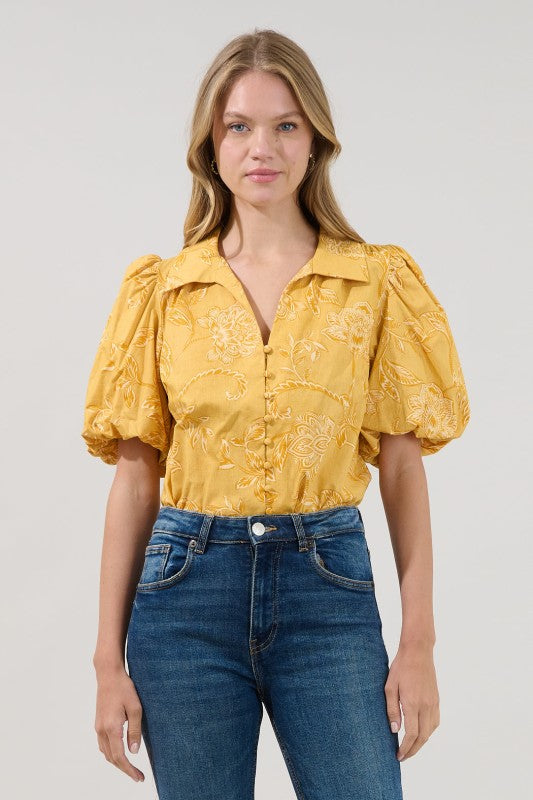 A woman modeling a yellow short-sleeve blouse with puff sleeves and tonal floral print, paired with blue jeans, against a light gray studio background.