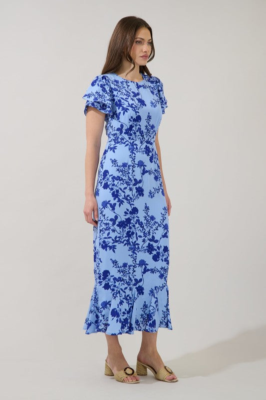 A woman modeling a light blue midi dress with a dark blue floral print, ruffled short sleeves, and a round neckline, standing against a plain light gray studio background.