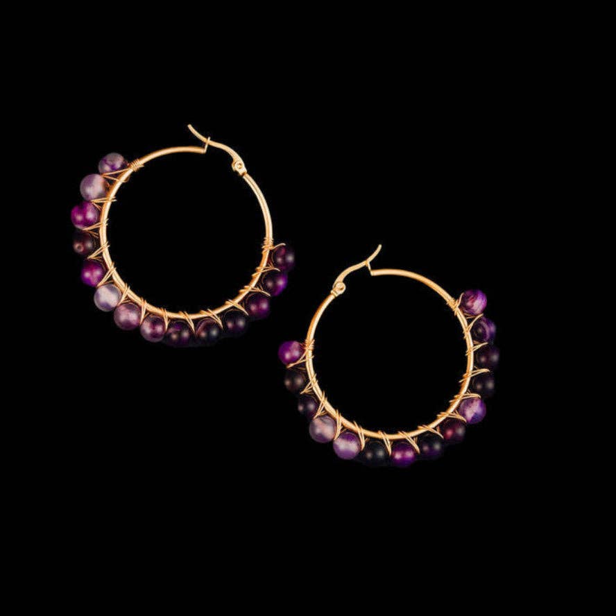 A pair of gold hoop earrings decorated with small purple and black beads, displayed against a solid black background.