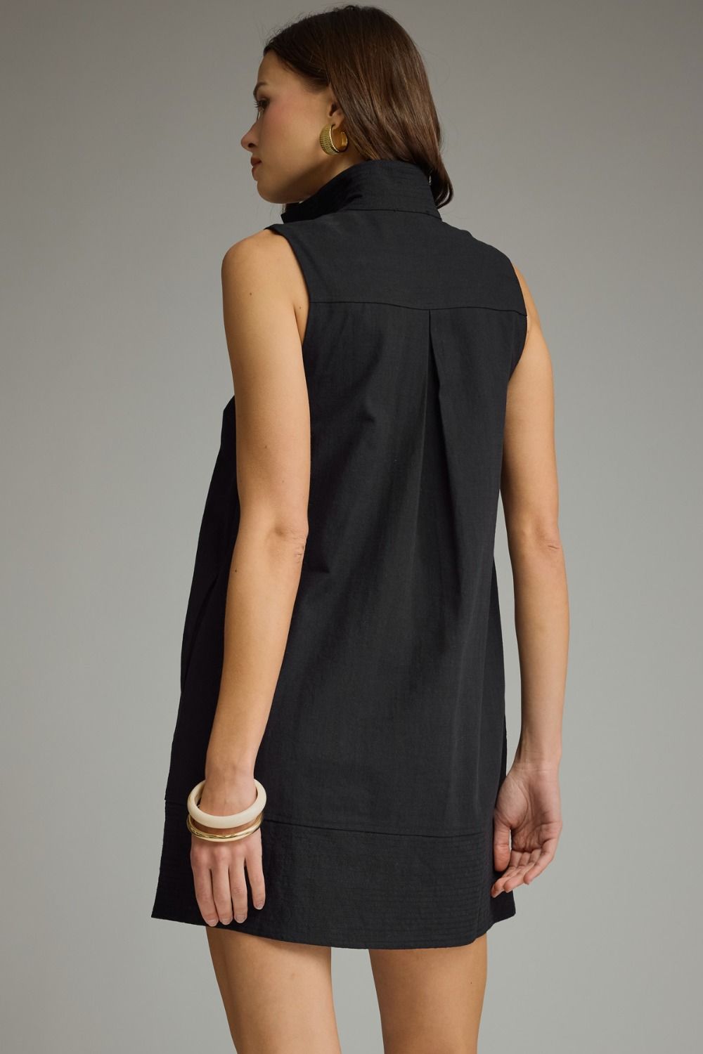 A woman shown from the back wearing a sleeveless black high-neck dress against a plain gray studio background.