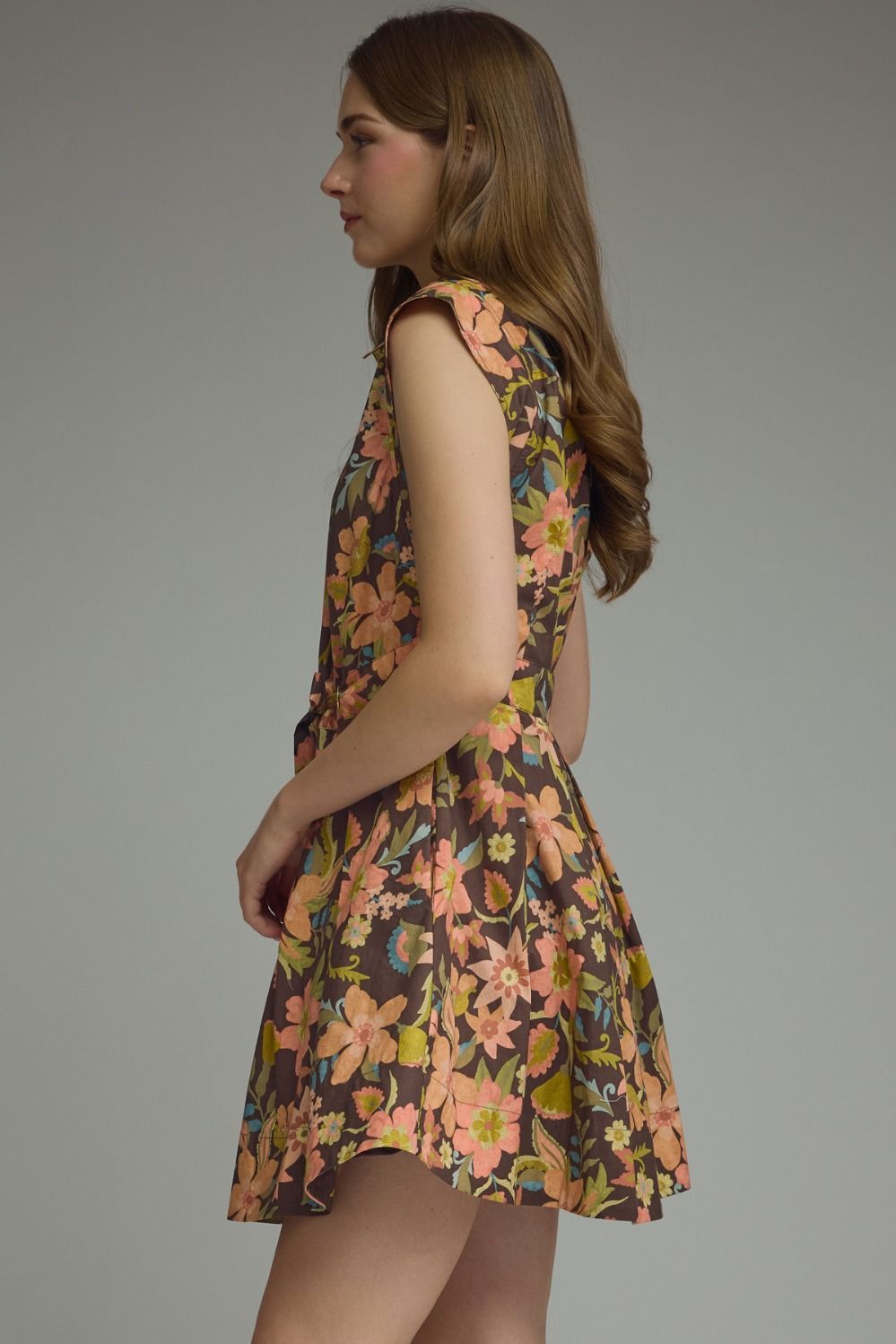 A woman wearing a sleeveless fit-and-flare dress with a colorful floral print on a brown base, photographed against a plain gray studio background.