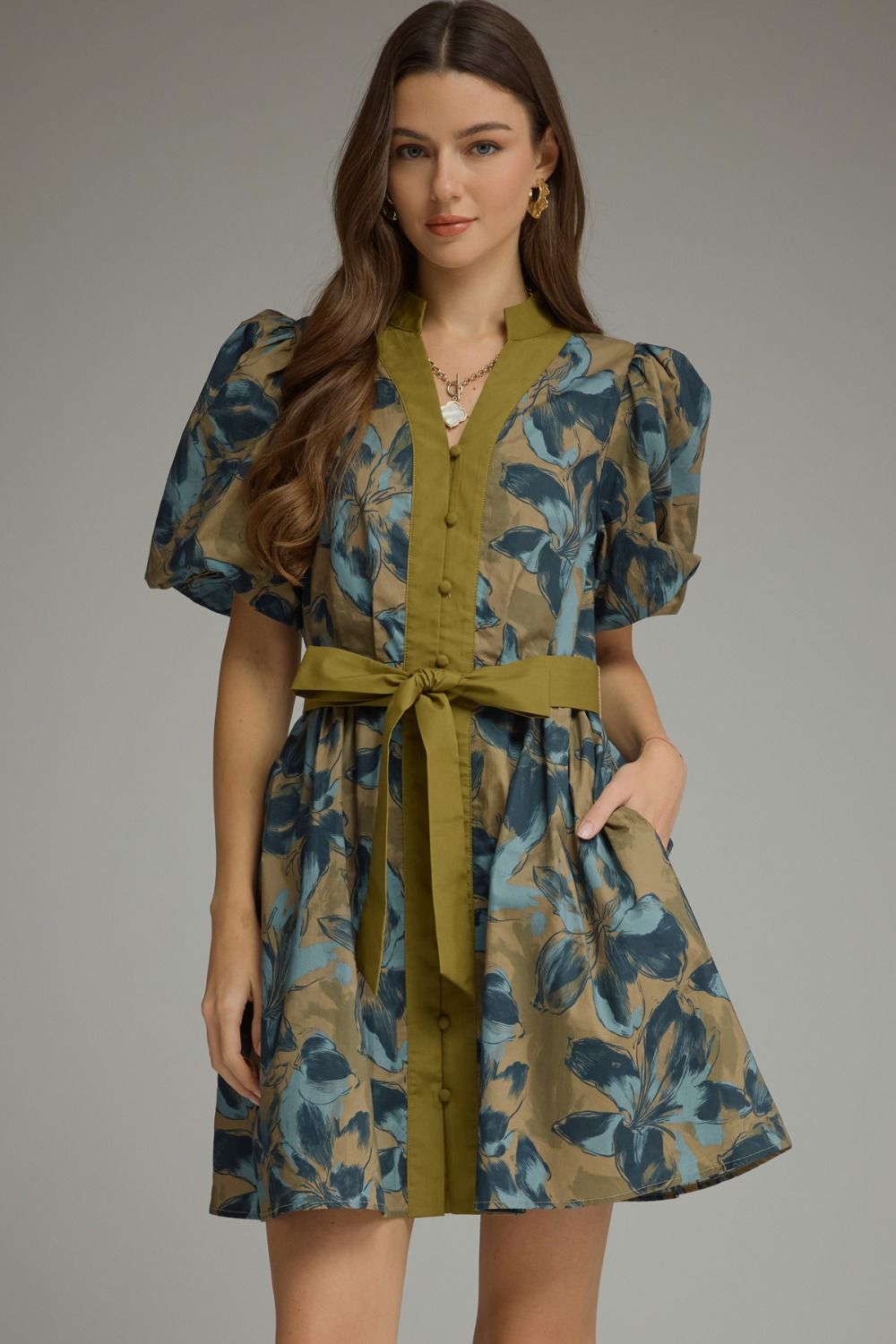A woman modeling a short puff-sleeve dress with a blue and olive green floral print, featuring an olive green button-front placket, matching waist tie belt, and side pockets, set against a solid gray studio background.