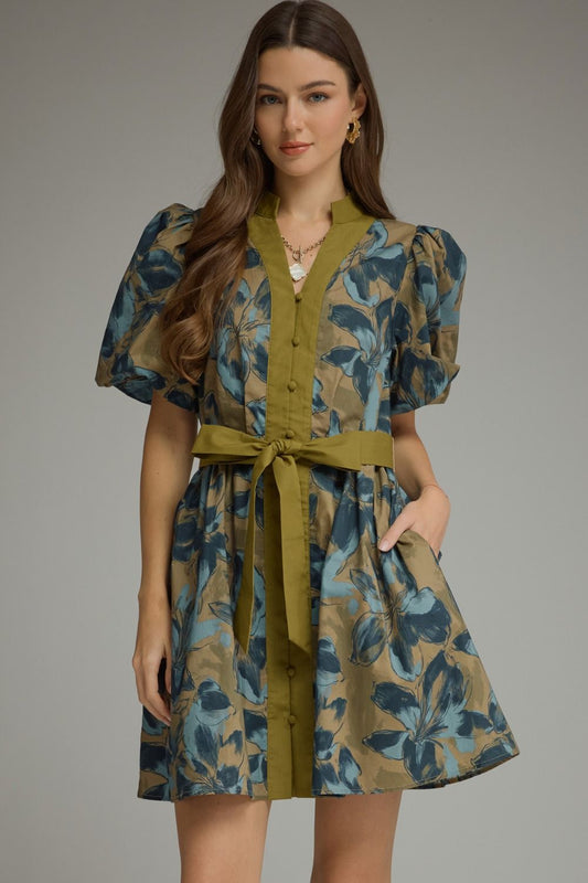 A woman modeling a short puff-sleeve dress with a blue and olive green floral print, featuring an olive green button-front placket, matching waist tie belt, and side pockets, set against a solid gray studio background.