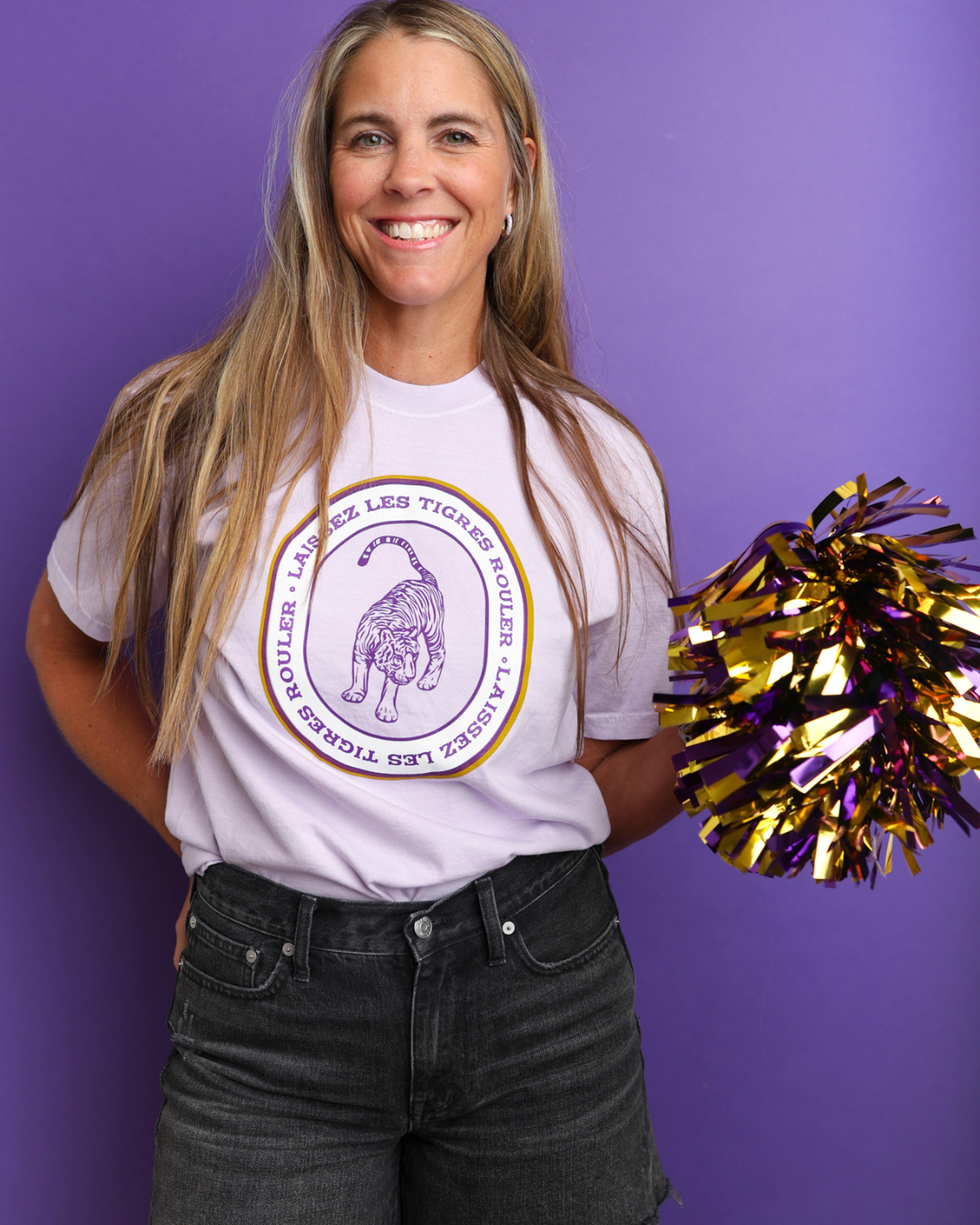 A smiling woman with long blonde hair stands against a solid purple background, wearing a lavender graphic t-shirt with a purple and gold tiger mascot design. She is holding a gold and purple pom-pom.
