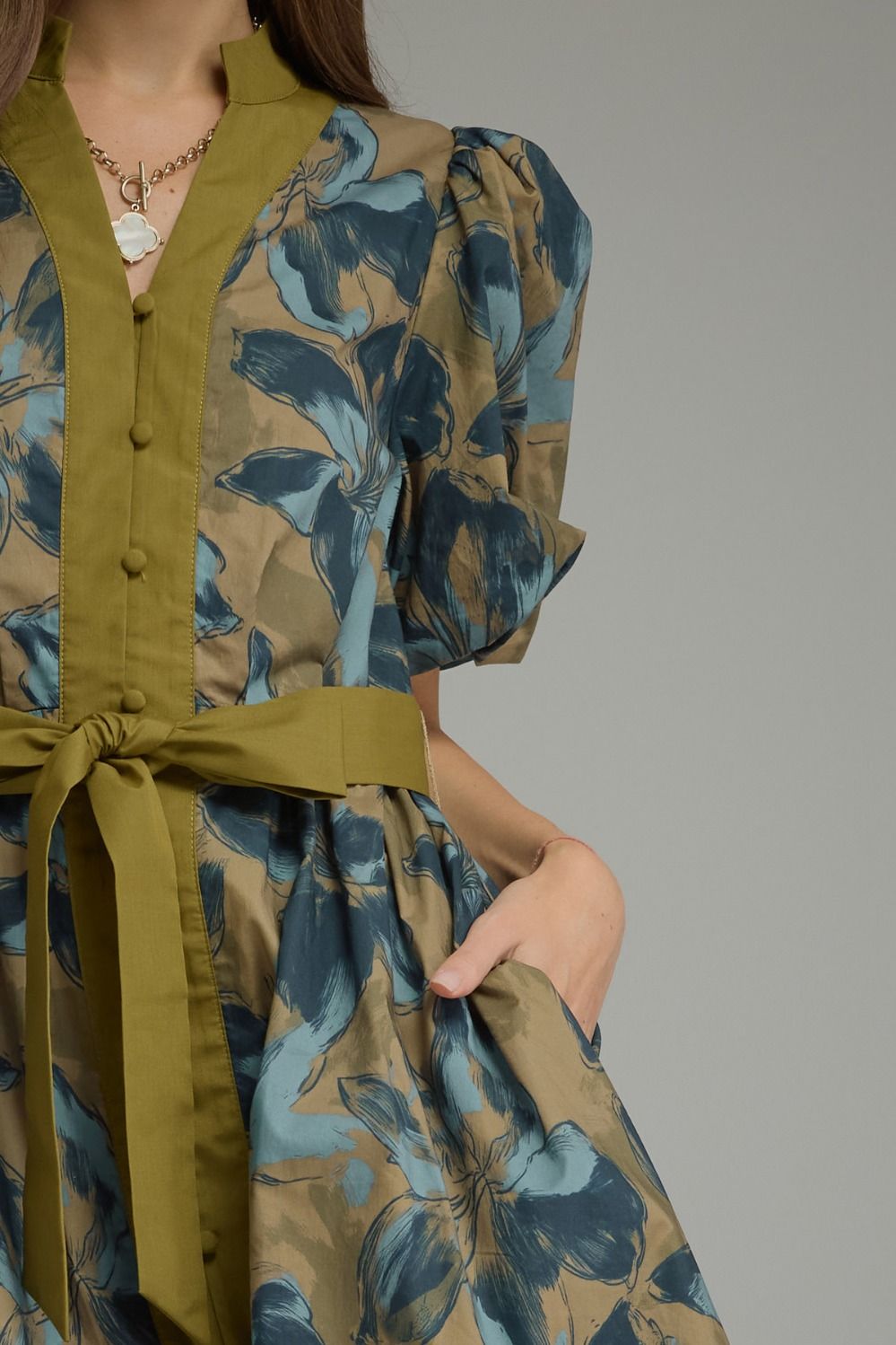 A close-up of a woman wearing a short-sleeved dress with a muted olive green collar, button placket, and matching waist tie, paired with a blue and brown floral print. The model is shown from the shoulders to the hips, with one hand in a pocket, against a solid gray background.