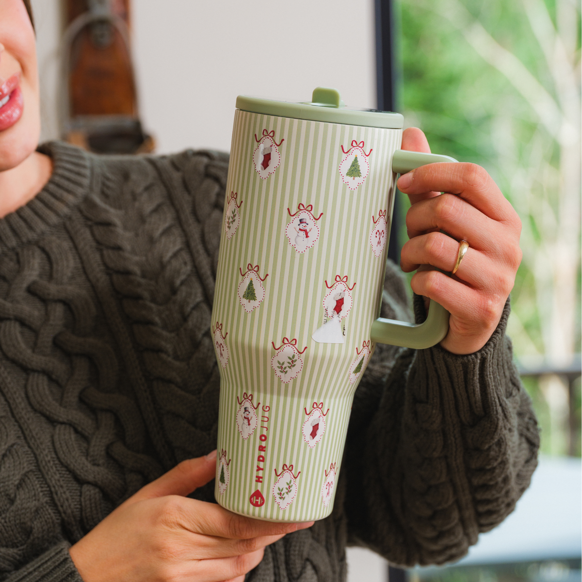 “Person holding Deck the Halls HydroJug tumbler with green stripes, Christmas motifs, and a green handle.”
