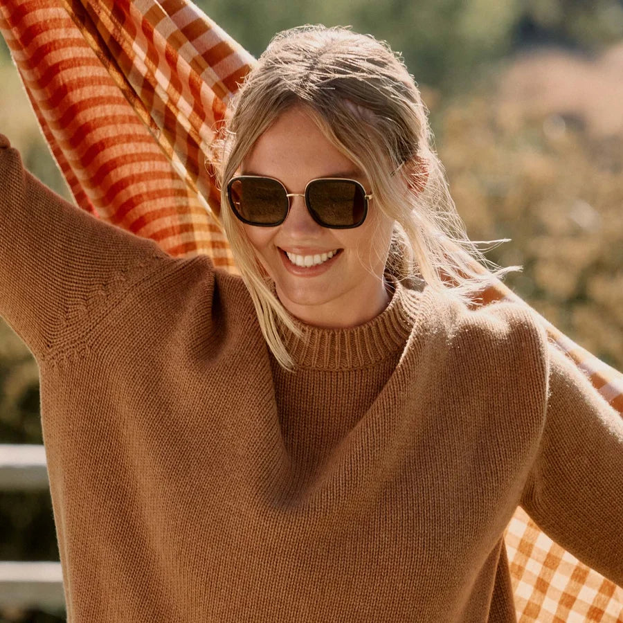 A woman wearing a warm brown ribbed knit sweater and dark sunglasses, smiling outdoors while holding an orange and white gingham blanket overhead.
