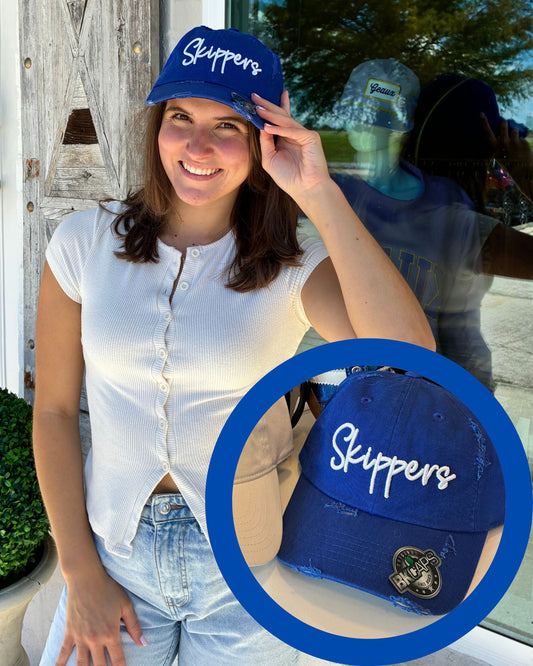 A woman wearing a blue baseball cap with 'Skippers' embroidered in white, shown in a lifestyle setting with a circular inset close-up of the cap. The cap has a distressed texture and a small embroidered patch on the side.