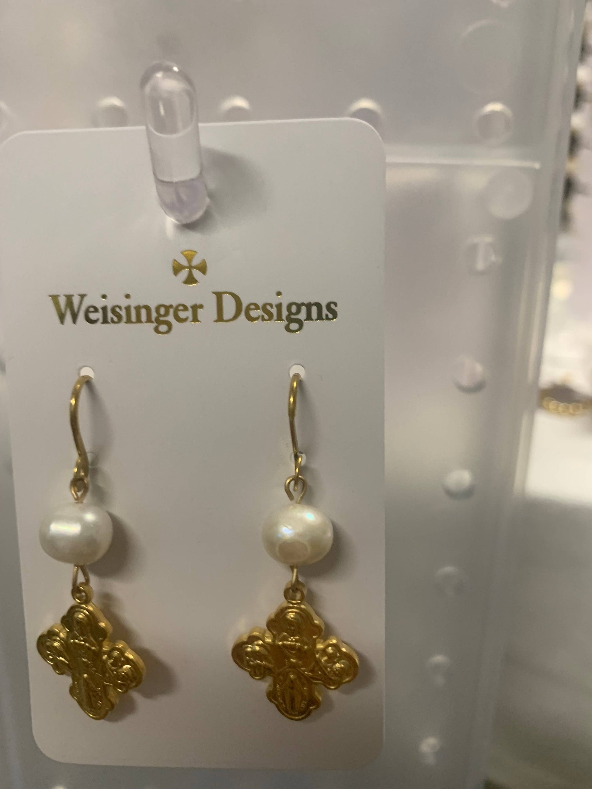 A pair of gold and pearl drop earrings by Weisinger Designs, featuring a round white pearl, a gold hook, and an ornate gold cross charm, displayed on a white card.