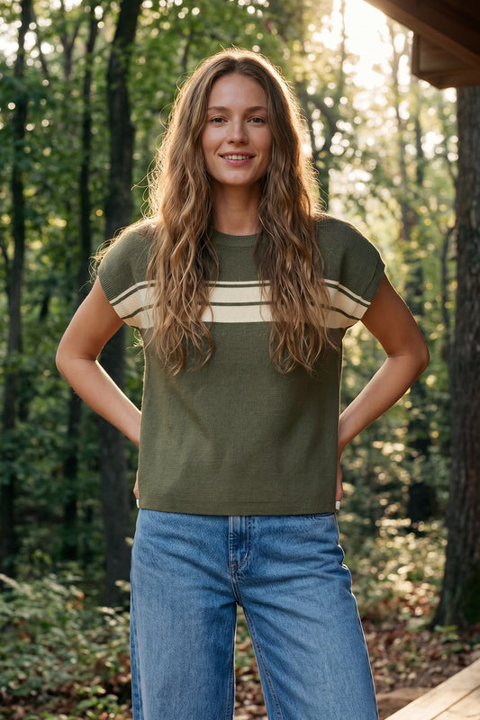 A woman standing outdoors in a wooded setting wearing an olive green short-sleeve knit top with beige horizontal stripes across the chest and blue jeans.