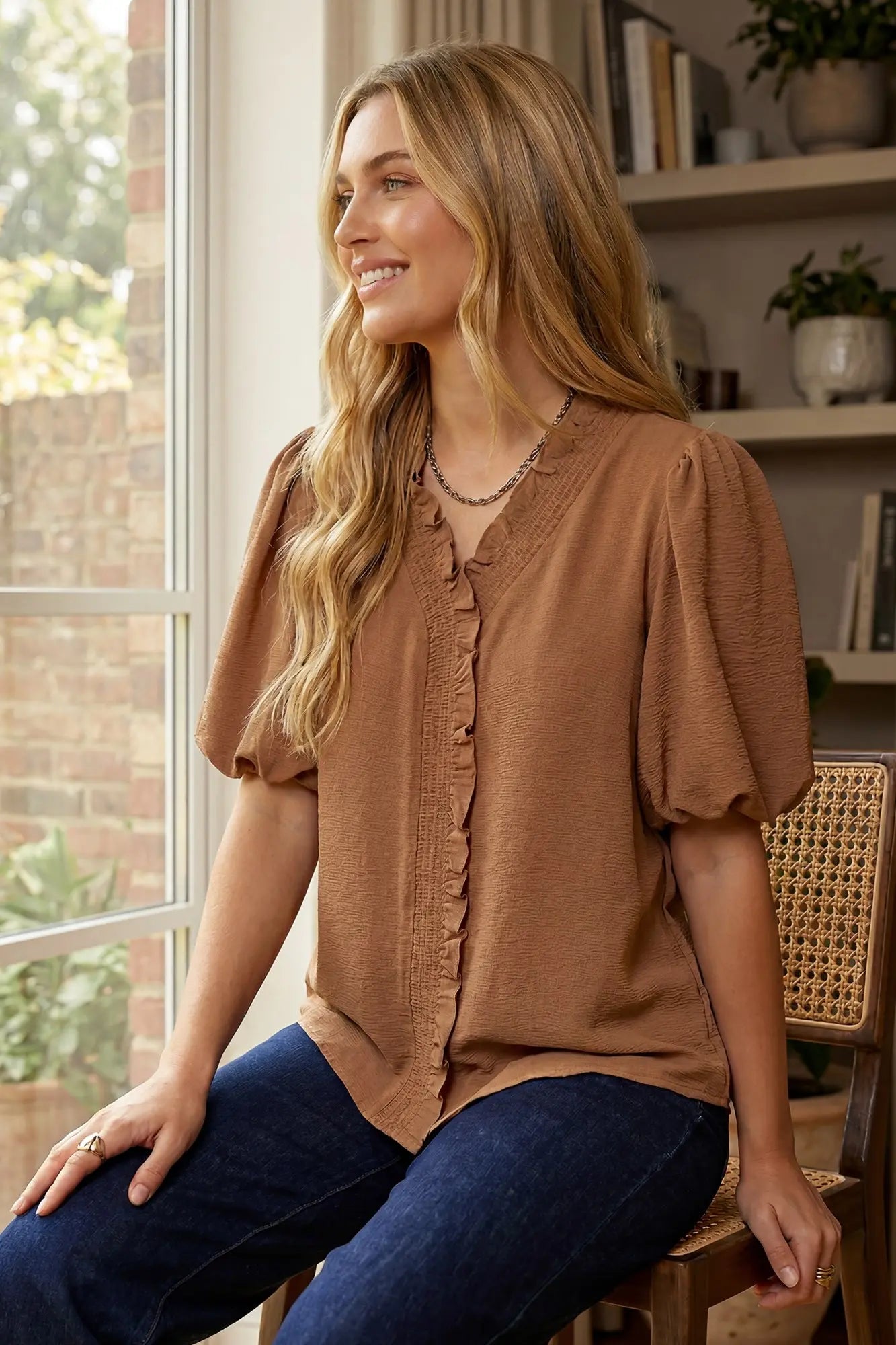 Woman sitting on a chair wearing a brown blouse and blue jeans, smiling.