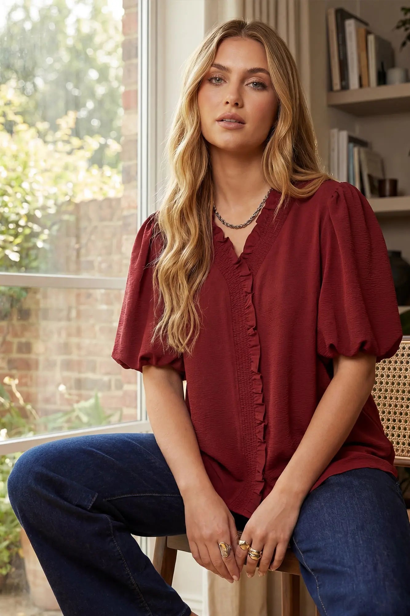 Woman wearing a wine blouse and blue jeans sitting indoors with a window and bookshelf in the background.