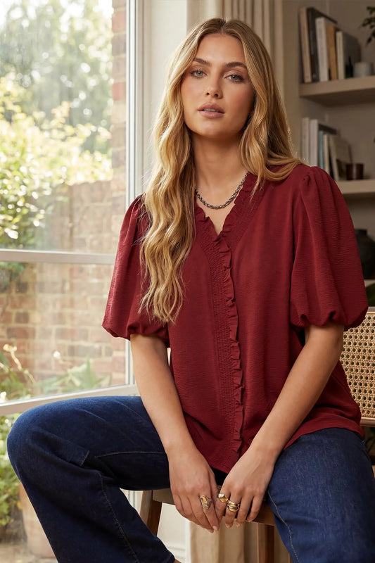 Woman wearing a wine blouse and blue jeans sitting indoors with a window and bookshelf in the background.