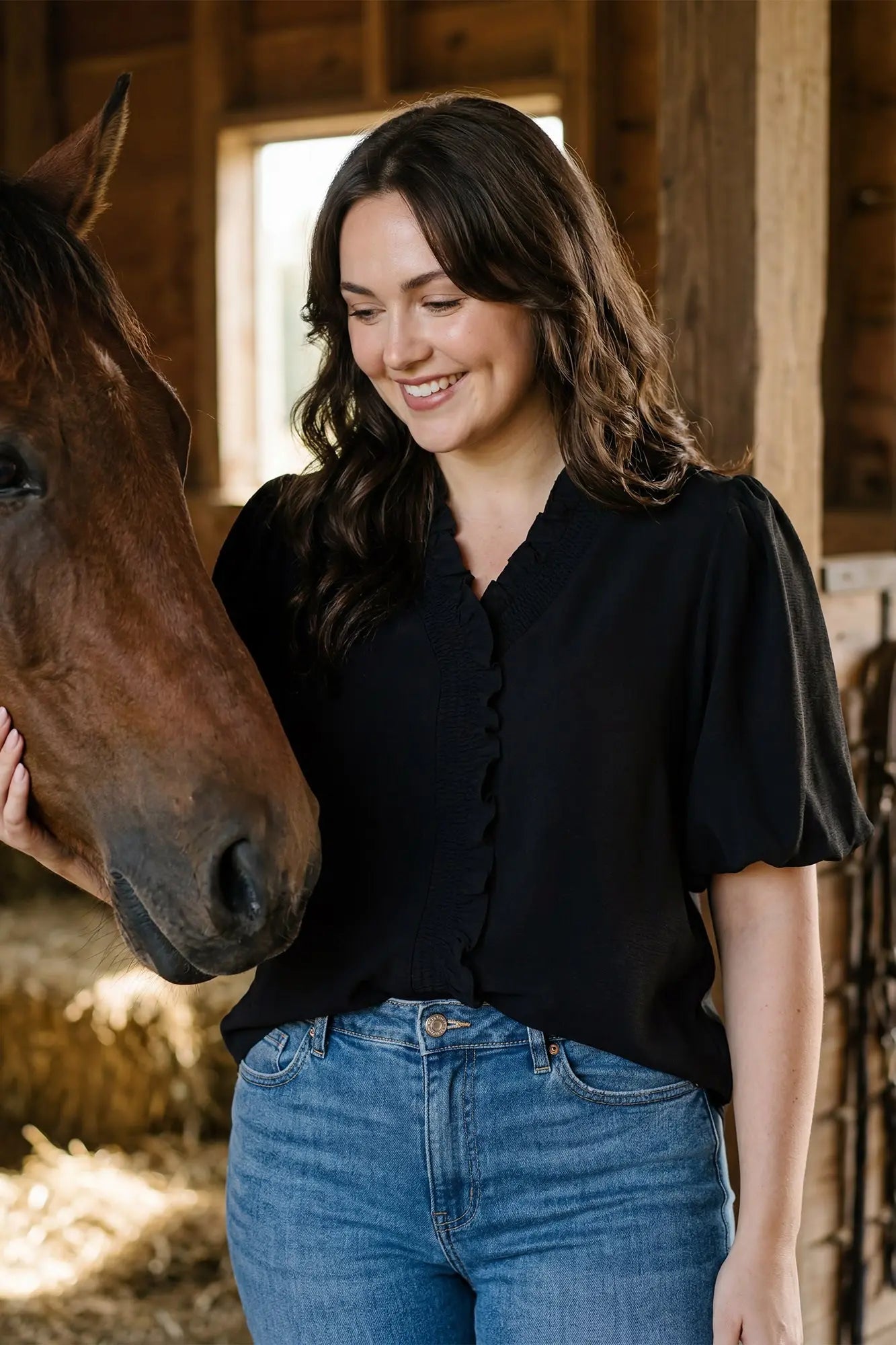 A woman wearing a black short-sleeve blouse with a scalloped ruffle trim and blue jeans, standing in a barn next to a brown horse.
