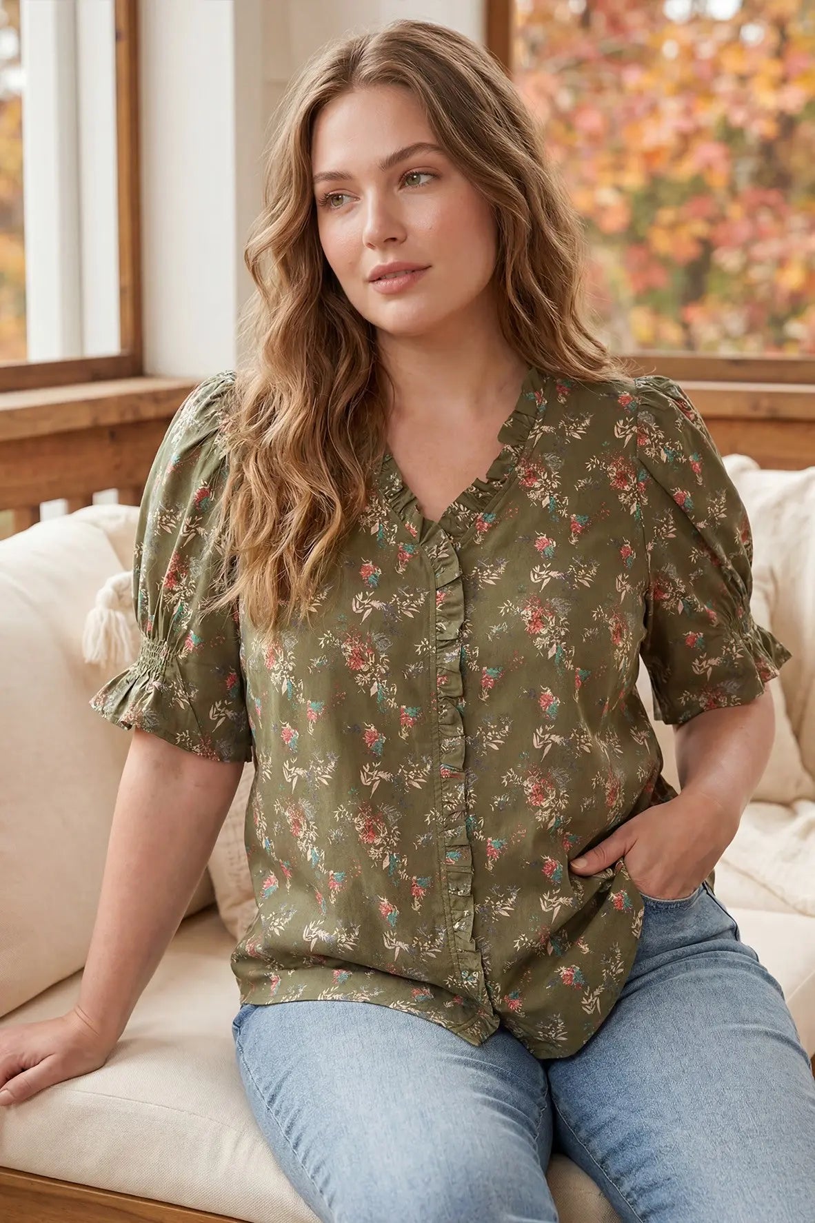 A woman wearing a short-sleeve olive green blouse with a delicate pink and red floral print, paired with light blue jeans, sitting on a cream-colored couch.
