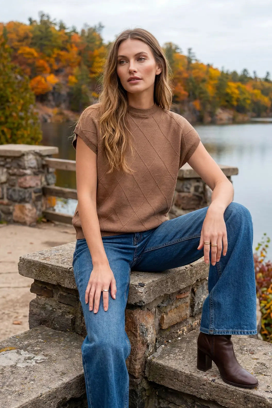A woman sits on a stone ledge outdoors by a lake, wearing a brown short-sleeve knit mock-neck top, blue jeans, and brown ankle boots. Trees with orange and yellow autumn foliage are visible in the background.