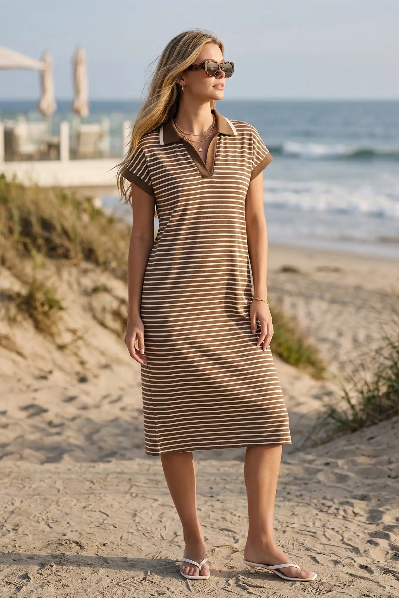 Woman in a mocha and off white striped dress standing on a sandy beach with ocean in the background