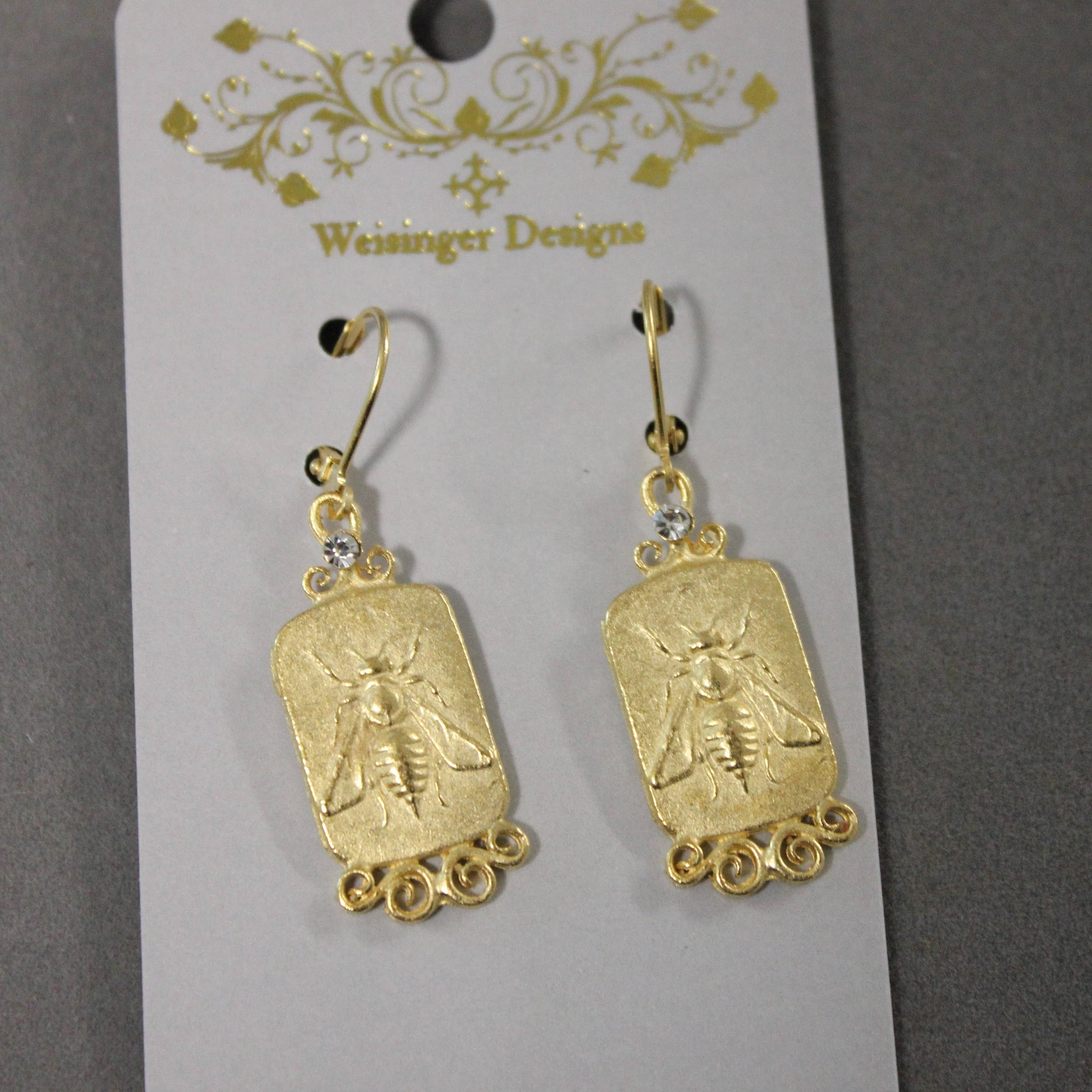 A pair of gold-toned drop earrings with rectangular bee-shaped charms and small clear rhinestones, hanging from a white display card printed with the brand name 'Weisinger Designs'.