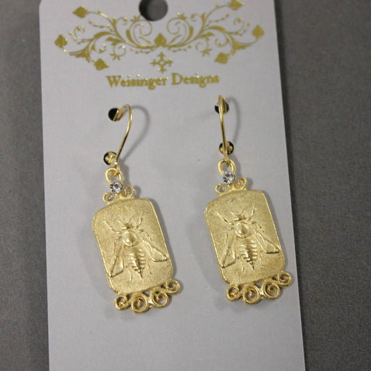 A pair of gold-toned drop earrings with rectangular bee-shaped charms and small clear rhinestones, hanging from a white display card printed with the brand name 'Weisinger Designs'.